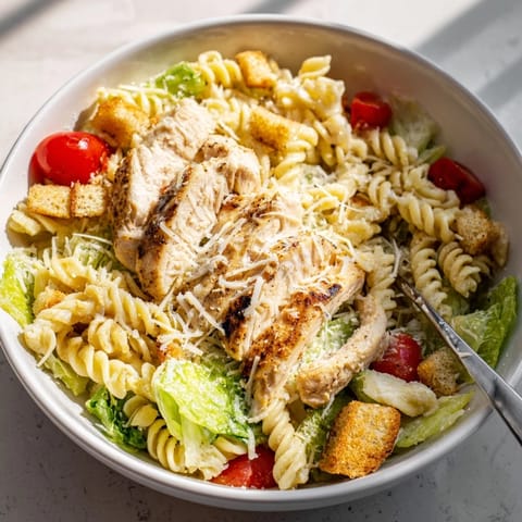 Caesar Pasta Chicken Bowl topped with garlic croutons, shaved Parmesan, and halved cherry tomatoes served on a white plate.  